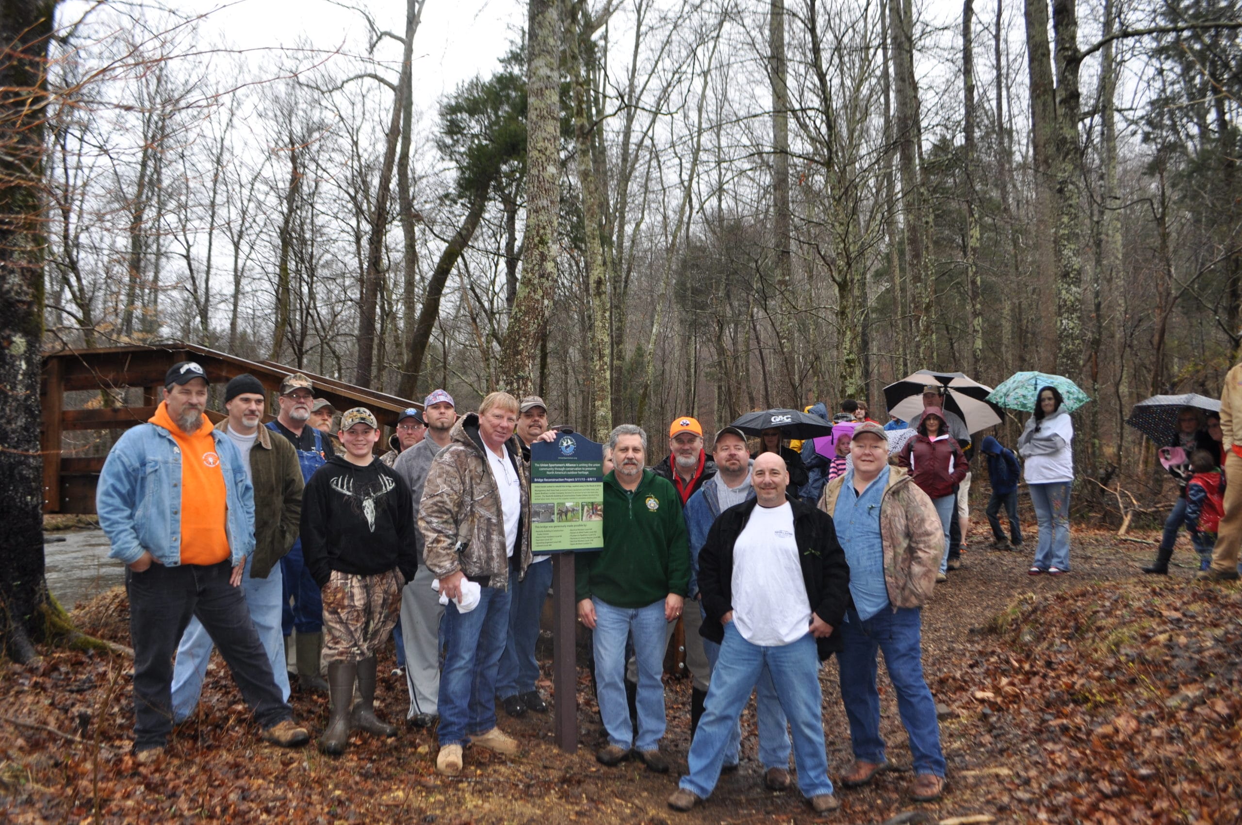 Tradesmen and women from the Nashville Building and Construction Trades who volunteered their time and talents to rebuild a bridge in the heart of Montgomery Bell State Park stand beside a permanent sign installed to recognize their contribution.